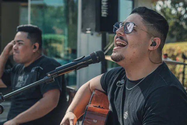 Cantor com violão e microfone durante uma apresentação, com um segundo músico ao fundo Cantor com violão e microfone durante uma apresentação, com um segundo músico ao fundo